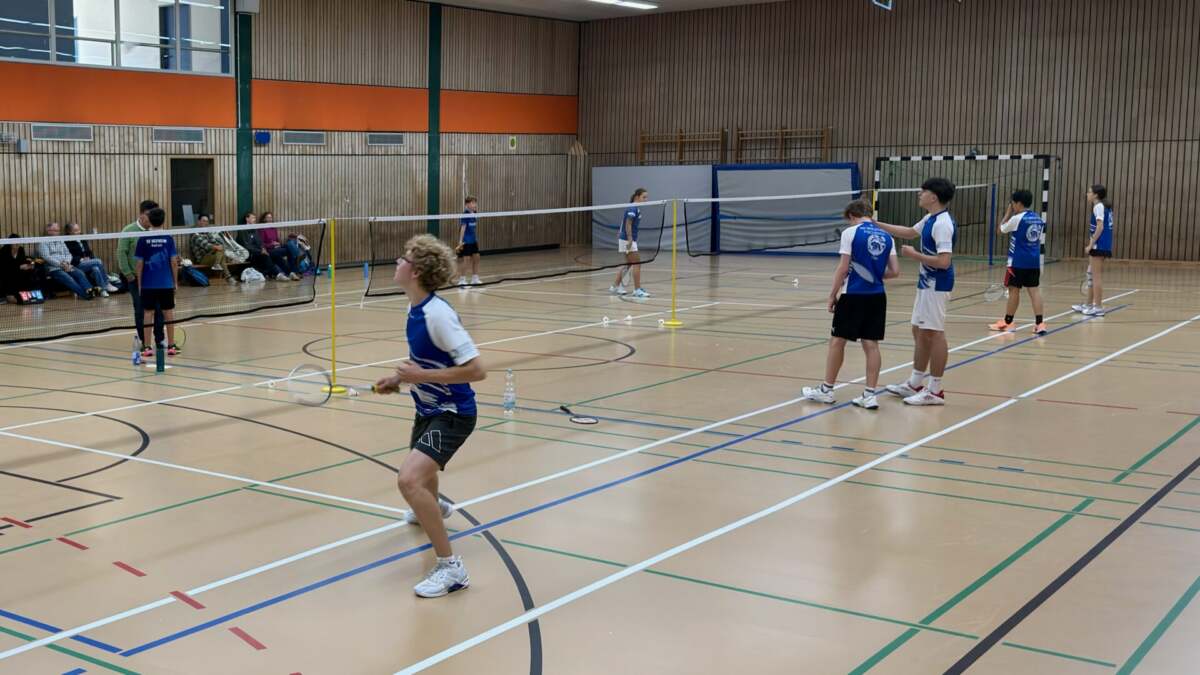 Badmintonspiel in der Comenius Halle: Ein Spieler in blau-weißem Trikot steht im Vordergrund am Netz, dahinter laufen weitere Spiele auf mehreren Feldern, am Rand sitzen Zuschauer